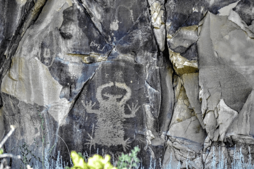 Petroglyph of a creature with outstretched arms on a rocky surface, surrounded by natural textures and colors.
