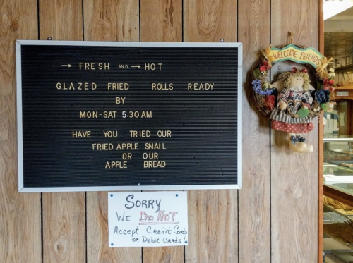 A sign on a wooden wall advertising fresh glazed fried rolls, apple snail, and apple bread; notes no credit/debit cards accepted.