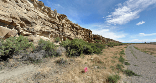 A rocky cliffside with sparse vegetation and a dirt path, under a clear blue sky with scattered clouds.