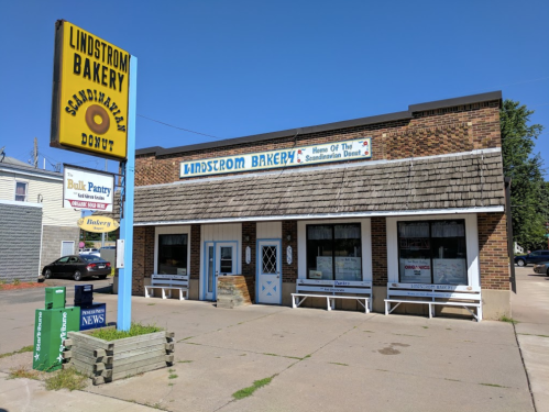 A brick bakery with a sign reading "Lindstrom Bakery" and "Home of the Scandinavian Donut," featuring outdoor seating.