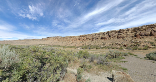 A wide landscape featuring rocky cliffs, sparse vegetation, and a clear blue sky with scattered clouds.