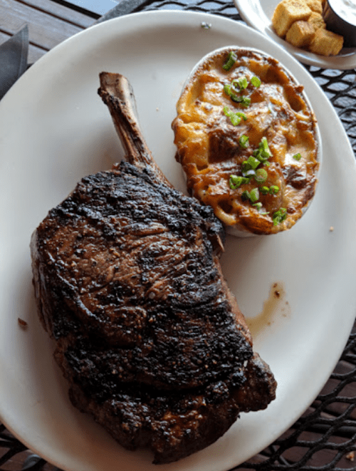 A large, grilled ribeye steak served with a cheesy potato dish and garnished with green onions on a white plate.
