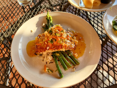A plate featuring grilled salmon on rice, topped with vegetables and served with asparagus, all drizzled in sauce.
