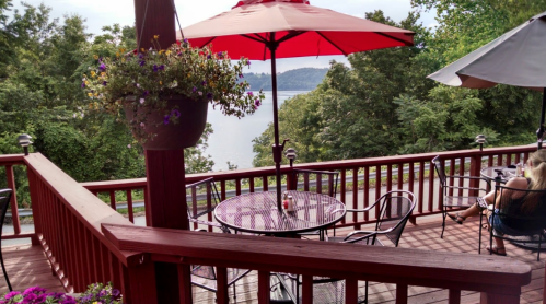 A scenic outdoor deck with tables, red umbrellas, and a view of a lake surrounded by trees.