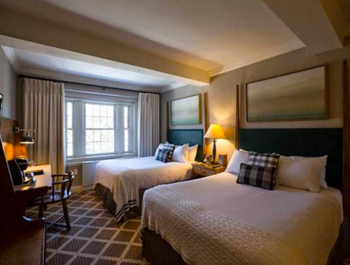 Cozy hotel room with two beds, a desk, and a large window, featuring warm lighting and neutral decor.
