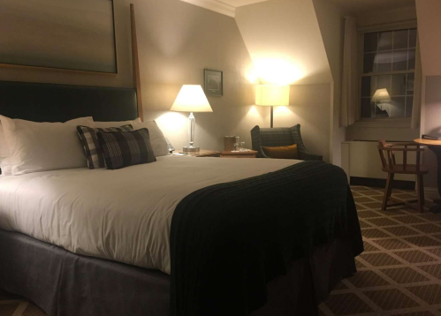 Cozy hotel room featuring a bed, lamps, a chair, and a desk with a window in the background. Warm, inviting atmosphere.