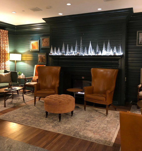 Cozy living room with two brown armchairs, a round ottoman, and a decorative shelf of illuminated trees.