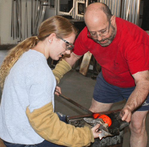 A man teaches a young woman glassblowing techniques in a workshop, both focused on the task at hand.