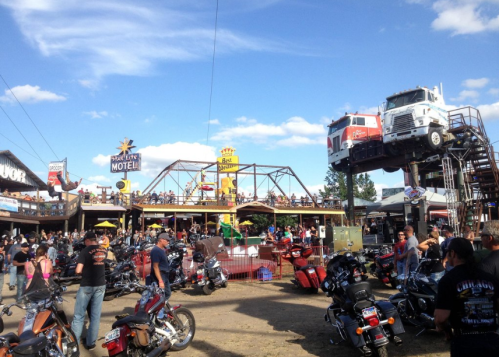 A lively outdoor motorcycle rally with crowds, bikes, and a large truck display against a blue sky.