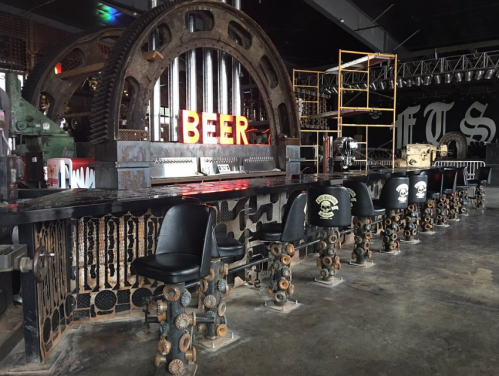 A unique bar with industrial decor, featuring a "BEER" sign and metal stools along a black countertop.