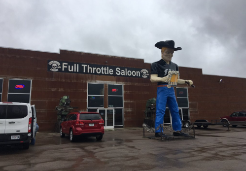 A large cowboy statue stands outside the Full Throttle Saloon, with vehicles parked in front and a cloudy sky above.