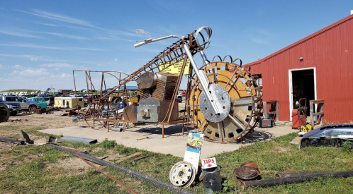 A large, artistic motorcycle sculpture made from metal parts, displayed outside a red barn in a grassy area.
