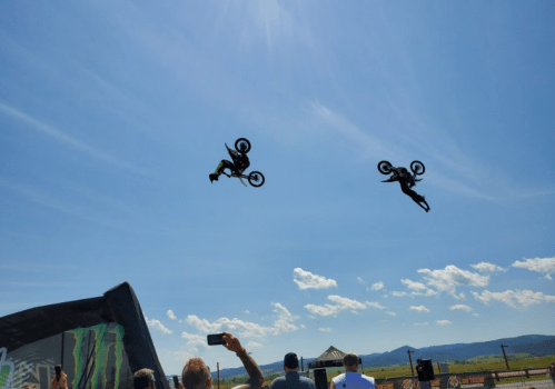 Two motocross riders performing flips in mid-air against a clear blue sky, with spectators watching below.