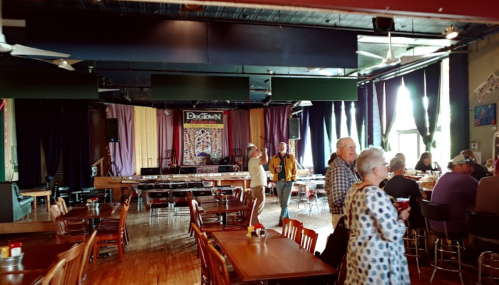 A lively restaurant interior with tables, a stage, and patrons enjoying their time. Colorful decor and a welcoming atmosphere.