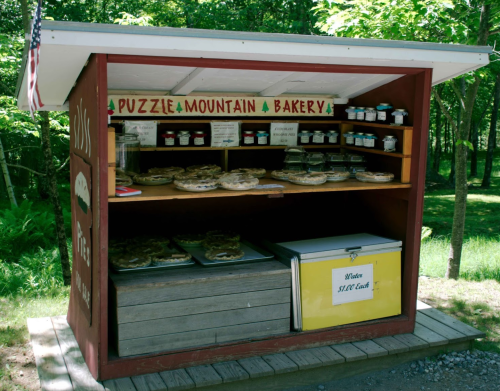 A rustic bakery stand with baked goods and jars of preserves, set in a wooded area.