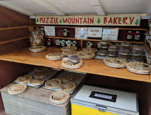 A rustic bakery shelf displaying various pies and baked goods, with a sign listing prices and offerings.
