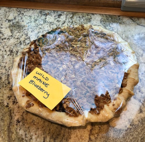A blueberry pie topped with granola, covered in plastic wrap, with a yellow sticky note labeled "WILD MAINE Blueberry."