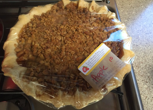 A freshly baked apple pie with a crumb topping, covered in plastic wrap, sitting on a countertop.