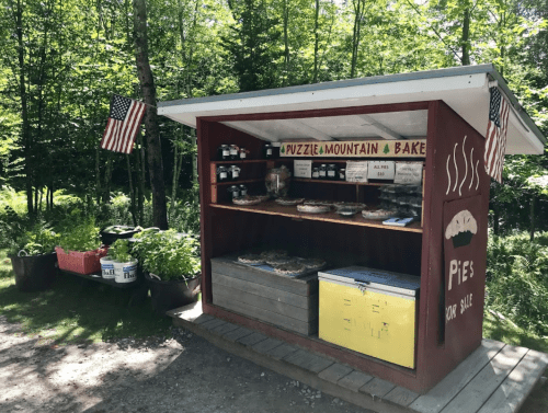 A rustic stand selling pies and jams, surrounded by greenery, with American flags displayed.