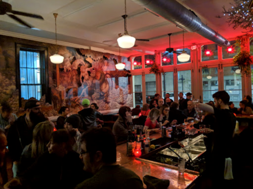 A lively bar scene with patrons socializing, colorful lighting, and a mural on the wall.
