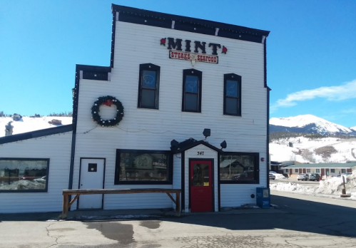 A white building with a red door and sign reading "MINT Steak & Seafood," surrounded by snow and mountains.