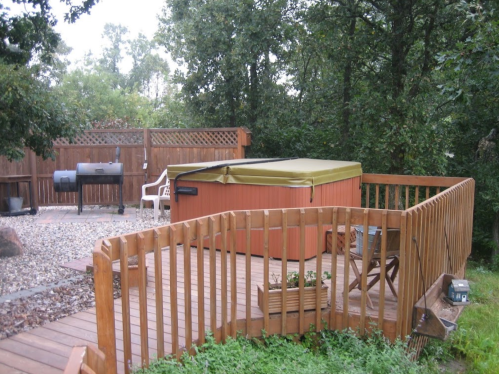 A wooden deck with a hot tub and grill, surrounded by trees and a fenced area.