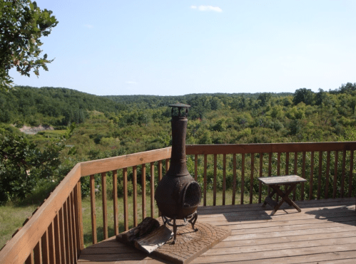 A wooden deck with a chiminea overlooks a lush green landscape under a clear blue sky.