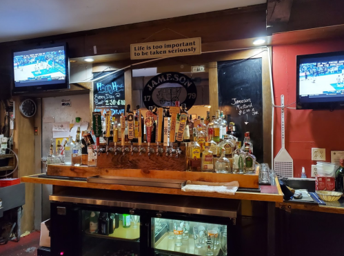 A bar with a wooden counter displaying various beer taps, two TVs showing sports, and a sign about taking life seriously.