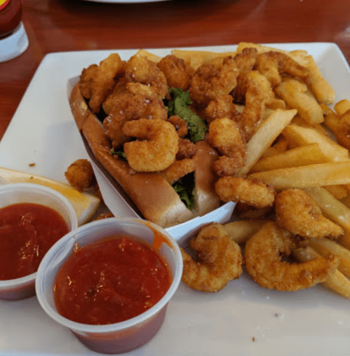 A plate with a shrimp sandwich, crispy fried shrimp, and a side of French fries, accompanied by two cups of dipping sauce.
