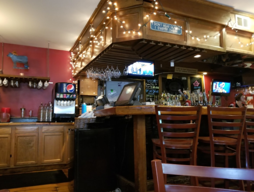 A cozy bar with wooden decor, string lights, a cash register, and TVs displaying drinks and menus.