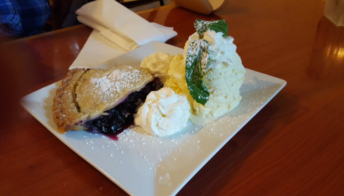 A slice of blueberry pie with whipped cream and a scoop of vanilla ice cream, garnished with mint leaves on a plate.