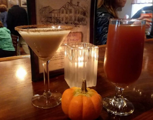 Two cocktails on a bar: a creamy drink in a martini glass and a red drink in a tall glass, with a small pumpkin and candle.