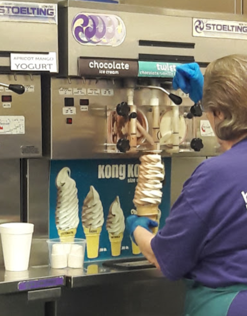 A person in a purple shirt serves a large swirl of chocolate ice cream from a machine, with cups nearby.