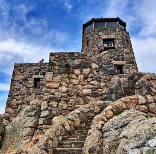 A stone tower with a viewing platform, surrounded by rocky terrain and a blue sky with clouds.