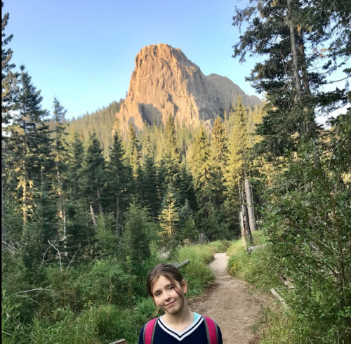 A girl stands on a forest path with a rocky mountain in the background, surrounded by tall trees and greenery.