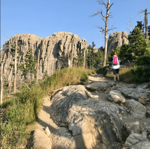 A hiker with a pink backpack walks along a rocky trail surrounded by trees and towering cliffs under a clear blue sky.