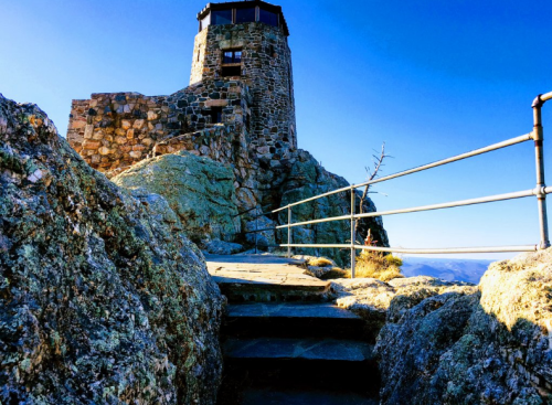 Stone tower on a rocky hilltop with a clear blue sky, featuring a railing and steps leading up to it.
