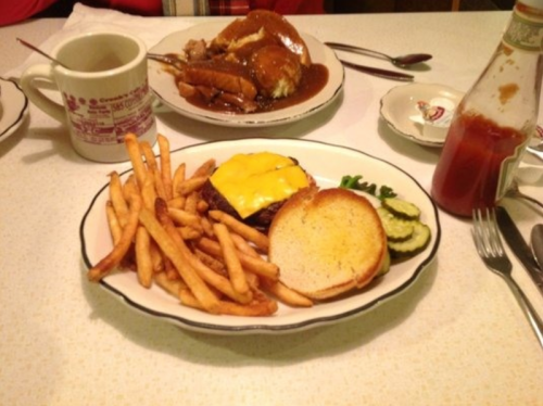 A plate with a cheeseburger, fries, pickles, and a side of ketchup, with a cup of coffee in the background.