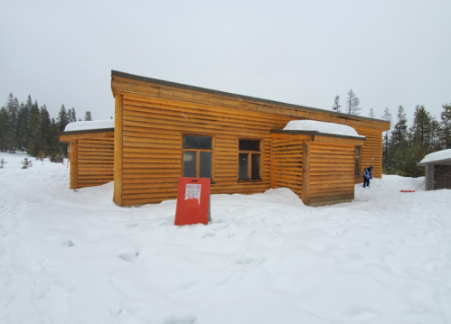 A wooden cabin in a snowy landscape, surrounded by trees, with a red sign in front and a person in the background.