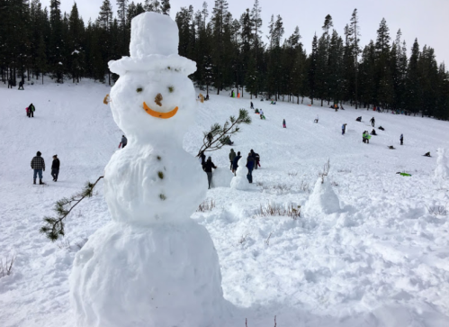 A cheerful snowman with a carrot nose stands in a snowy landscape, with people sledding in the background.