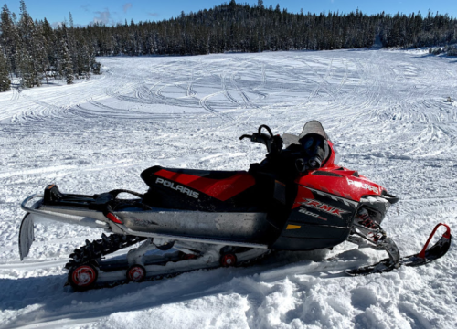 A red and black snowmobile parked on a snowy landscape with tire tracks in the snow and trees in the background.