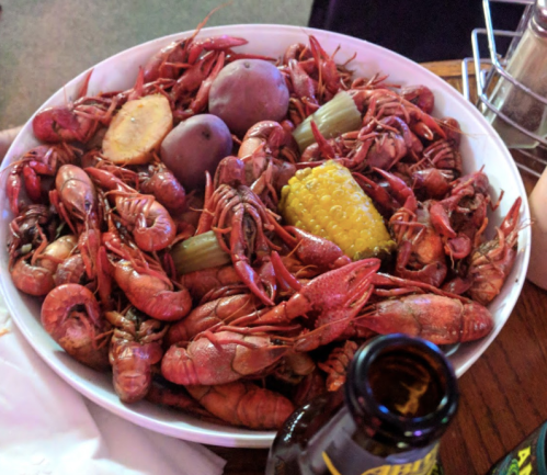 A large bowl filled with crawfish, potatoes, corn, and celery, with a bottle of drink beside it.