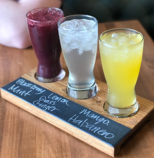 Three colorful drinks on a wooden tray: a dark berry drink, a clear soda, and a yellow mango beverage, labeled with flavors.