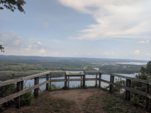 Scenic overlook with wooden railing, showcasing a river and lush green landscape under a partly cloudy sky.