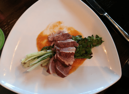 A plate with sliced duck breast, garnished with greens and a drizzle of sauce, served on a white dish.