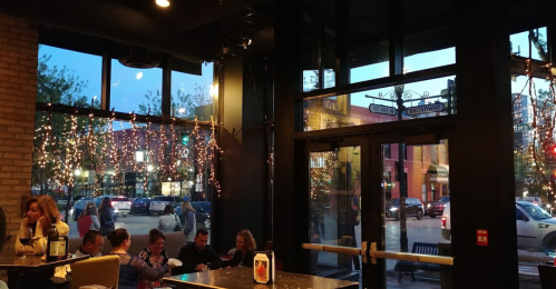 A cozy restaurant interior with string lights, large windows, and patrons enjoying their meals as evening falls outside.