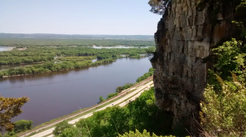 A scenic view of a river winding through lush greenery, with a rocky cliff in the foreground under a clear blue sky.