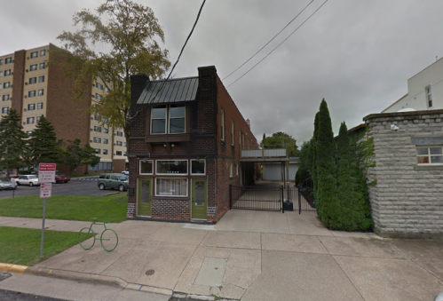 A two-story brick building with a green door, surrounded by trees and parked cars on a cloudy day.