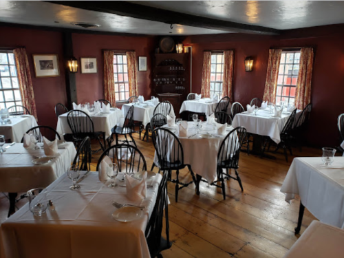 Cozy restaurant interior with wooden floors, white tablecloths, and warm lighting. Tables set for dining.