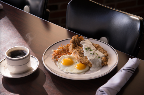 A plate with fried chicken, two sunny-side-up eggs, hash browns, and gravy, alongside a cup of steaming coffee.